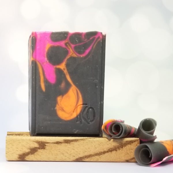 hot pink and orange swirls on a black soap bar. Shown on a wooden soap dish with soap curls.